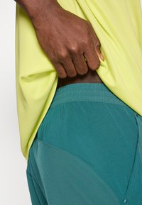 Κίτρινο πουκάμισο με κοντά μανίκια και υφή υφάσματος, συνδυασμένο με τυρκουάζ σορτς με ελαστική μέση και πλαϊνές τσέπες.