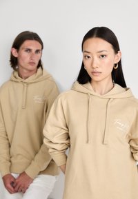 Två personer bär beige huvtröjor med en framficka och dragsko. Huvtröjorna har en mjuk textur och subtil logotypdetalj.