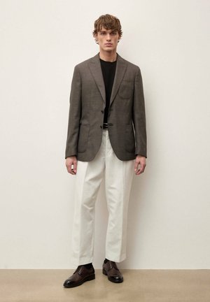 Jeune homme portant un blazer marron, une chemise noire, un pantalon blanc à jambes larges et des chaussures habillées marron foncé, debout contre un mur uni.