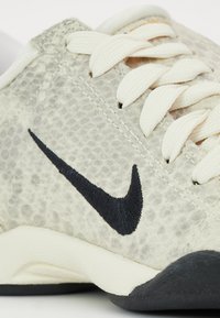 Zapatilla con una parte superior texturizada de gris claro, logotipo de Nike en negro y cordones blancos; presenta una suela flexible de color crema con detalles en negro.