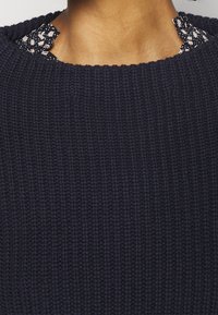 Granatowy sweter w robionym na drutach fasonie, z teksturowaną powierzchnią, z ozdobnym kołnierzykiem w czarno-białe wzory wystającym z dekoltu.