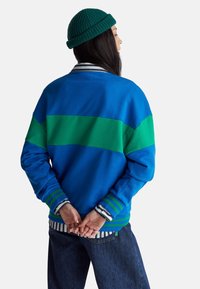 Felpa blu con strisce orizzontali verdi, polsini a costine e una camicia a strisce a strati sottostante. Indossata con jeans blu scuro e un berretto verde.