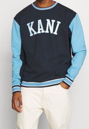 Homme portant un sweat-shirt à manches longues bleu marine et bleu clair avec le texte « KANI » sur la poitrine et un pantalon écru.