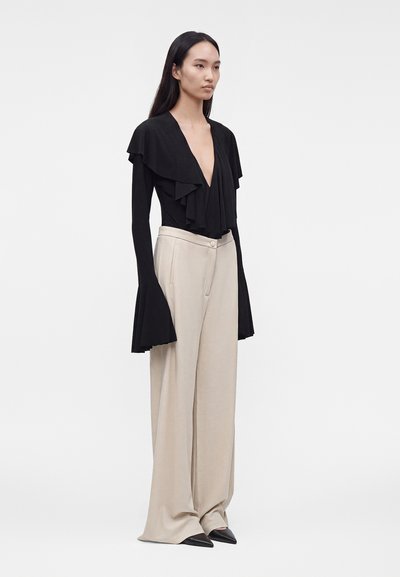 Femme aux longs cheveux foncés portant un top noir à volants avec un décolleté en V profond, des manches évasées et un pantalon beige large avec des chaussures noires pointues.