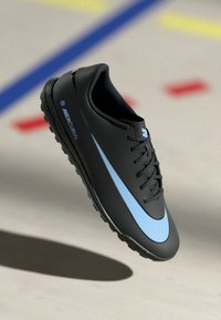 Must musta Nike Mercurial jalgpallijalats, millel on helesinine swoosh, mustad paelad ja tekstureeritud tald, kuvatud üle siseruumi kohtu põranda, millel on värvilised jooned.