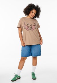 Beige Grafik-T-Shirt mit schwarzem und pinkem Flügel-Design, kombiniert mit dunkelblauen Jeansshorts und grünen Sneakers. Schlichter Hintergrund.