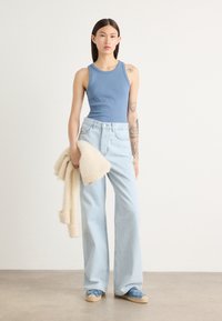 Lichtblauwe ribbel tanktop gecombineerd met hoge taille, wijde lichtdenim jeans. Houdt een crèmekleurige, zachte gebreide cardigan vast. Blauwe gestreepte espadrilles.