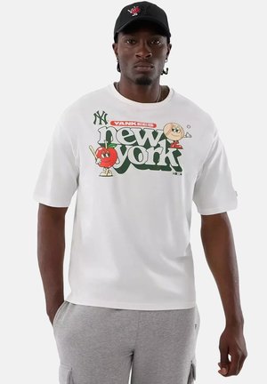 Homme portant un t-shirt blanc New York Yankees avec des personnages de pomme et de baseball en dessin animé, pantalon gris et casquette de baseball noire.