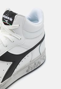 Vit hög sneaker av läder, med en svart sidoree, texturerad sulan och en vadderad blå foder vid kragen.