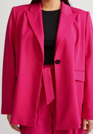 Mujer con un blazer ajustado de color rosa brillante y pantalones de talle alto a juego, con una camiseta negra de canalé debajo.