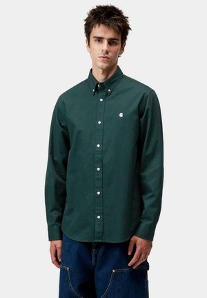Camisa de botones verde de manga larga con cuello, que presenta un pequeño logo en el pecho, combinada con pantalones anchos de mezclilla azules.