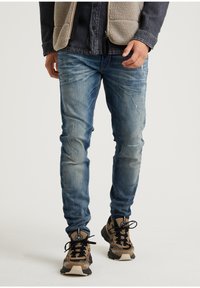 Vaaleansiniset, kulutetut farkut, joissa on slim fit -malli. Yhdistetty ruskeisiin kuvioituihin tennareihin. Tausta on neutraali ja huomaamaton.