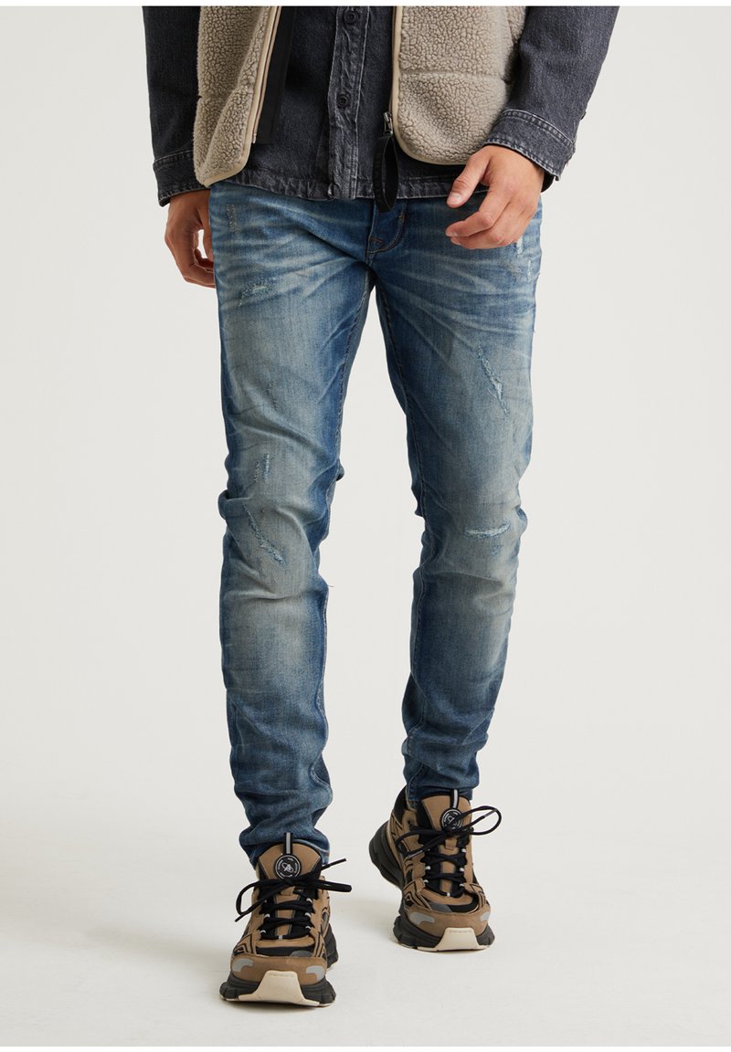 Vaaleansiniset, kulutetut farkut, joissa on slim fit -malli. Yhdistetty ruskeisiin kuvioituihin tennareihin. Tausta on neutraali ja huomaamaton.