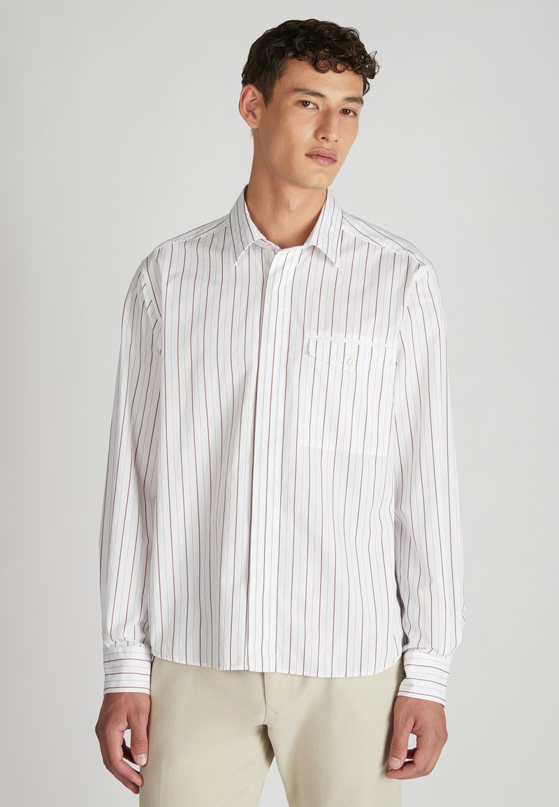 Camicia bianca a maniche lunghe con sottili righe verticali rosse, colletto button-down e taschino sul petto, abbinata a pantaloni beige. Texture liscia.