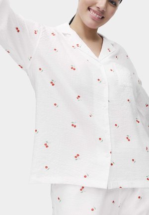 Lächende Person in weißen Knopfpajamas mit kleinen roten Kirschmustern, die einen Arm vor einem einfarbigen Hintergrund hebt.