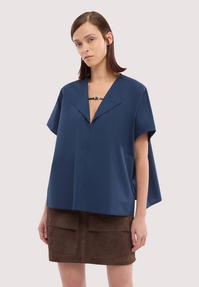 Kobieta stojąca, ubrana w luźną, granatową, krótkorękawową bluzkę oraz brązową zamszową spódnicę z dużymi przednimi kieszeniami na jednolitym tle.