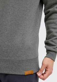 Sudadera gris hecha de un tejido suave con puños y dobladillo acanalados. Presenta una etiqueta de cuero marrón en la cintura. Ajuste elástico y cómodo.