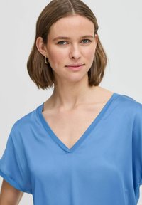 Helehall sinine V-kaelusega lühikeste varrukatega topp, mis on valmistatud siledast kangast. Omab lõdva lõikega disaini, kerget läiget ja puhta viimistlusega kaelust.