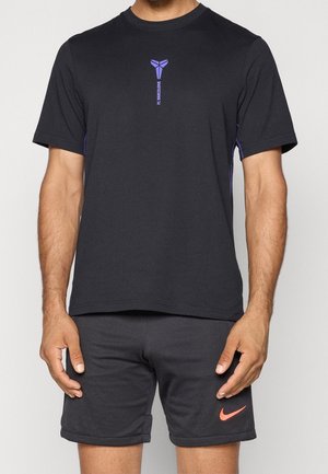 Mann trägt schwarzes FC Barcelona-T-Shirt mit lila Logo und schwarze Nike-Shorts mit orangefarbenem Swoosh, steht mit entspannten Armen.