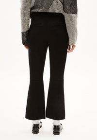 Schwarze, ausgestellte Hosen mit einer glatten Textur, ausgestattet mit zwei Gesäßtaschen und einem mittelhohen Bund. Kombiniert mit schwarzen Schuhen und weißen Socken.