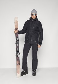 Chaqueta de esquí negra con cuello alto y cremalleras, pantalones de esquí negros, gorro gris, gafas de sol reflectantes, sosteniendo esquís con fijaciones.