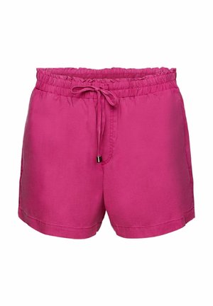 Femme aux longs cheveux ondulés portant une chemise à manches longues magenta et un short assorti, debout de dos, avec des sandales noires.