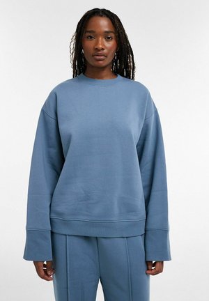 Vrouw met gevlochten haar draagt een losse blauwe sweater en bijpassende blauwe broek tegen een effen witte achtergrond.