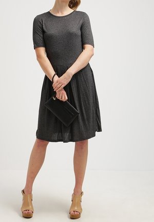 Grijze jurk met korte mouwen, een aansluitend lijfje en een wijd uitlopende rok, gecombineerd met een zwarte clutch en beige platformsandalen.