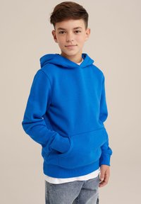 Blauer Hoodie aus weichem Material mit einer aufgesetzten Tasche und verstellbarem Kordelzug in der Kapuze, kombiniert mit hellen Jeans für einen lässigen Look.