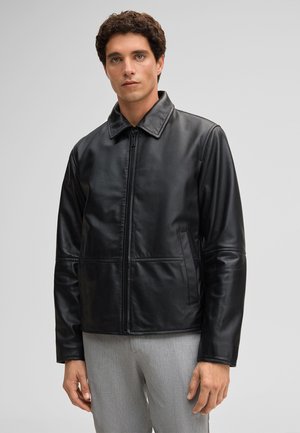 Mann mit kurzem, lockigem Haar steht frontal und trägt eine schwarze Lederjacke und eine hellgraue Hose vor einem schlichten, hellen Hintergrund.