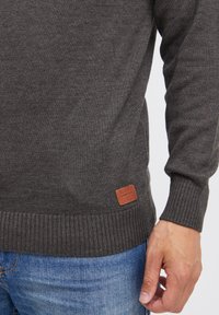 Mørkegrå strikket genser med ribbede ermer og kant, med et brunt skinnmerke med "BLEND" på den nedre siden. Denim jeans synlige.