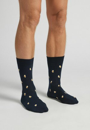 Benen in zwart enkelsokken met een patroon van kleine biermok-iconen, staand op een effen lichtgrijze ondergrond.