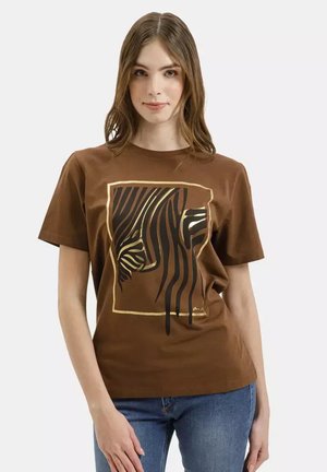 Kobieta o jasnobrązowych włosach ma na sobie brązowy T-shirt z abstrakcyjnym wzorem falistych czarno-złotych pasków, w połączeniu z niebieskimi dżinsami, stojąca na białym tle.