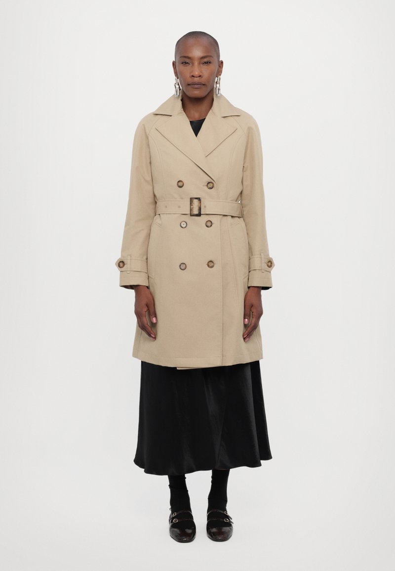 Beige trench coat with double-breasted design, belted waist, and large buttons. Worn over a black satin skirt and dark socks.