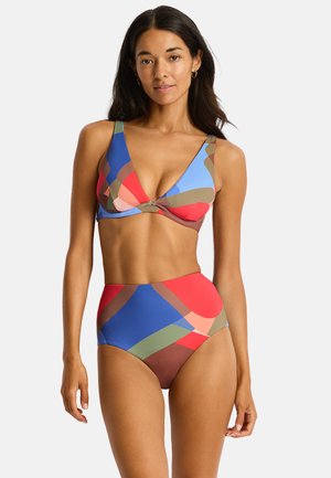 Kobieta ubrana w kolorowe bikini z abstrakcyjnym wzorem, z topem z dekoltem w serek i majtkami z wysokim stanem, stojąca na jednolitym jasnym tle.