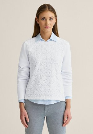 Kvinde med glat hår iført hvid sweater med blondevæv over en lyseblå skjorte med krave og grå bukser med smalle striber, stående mod en neutral baggrund.