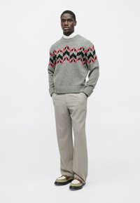 Γκρίζα πλεκτή ζακέτα με κόκκινο, μαύρο και μπορντό γεωμετρικό σχέδιο· συνδυάζεται με ανοιχτό γκρι παντελόνι και υποδήματα δύο τόνων, με φόντο απλού χρώματος.