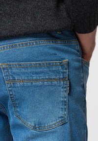 Jean denim bleu avec couture orange, comprenant une poche arrière et une étiquette de marque sur le côté. Texture lisse, design décontracté.