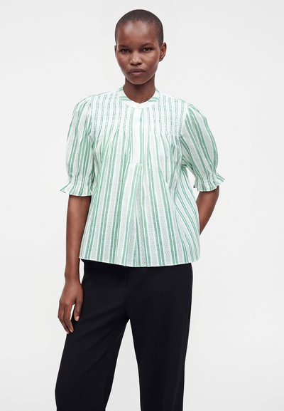Femme portant un chemisier à rayures verticales vertes et blanches avec des manches bouffantes et un pantalon noir, debout contre un fond blanc uni.