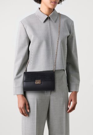 Une femme porte un costume à carreaux noir et blanc avec un sac bandoulière en cuir noir, orné d'un rabat à texture crocodile et d'une sangle chaîne dorée.