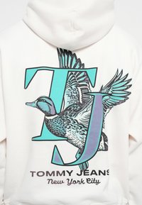 Hvit hettegenser med et stort grafisk motiv av enand ved siden av bokstavene "TJ" i teal og lilla. Teksten sier "TOMMY JEANS New York City."