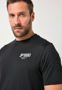 Czarny bawełniany t-shirt z krótkimi rękawami, okrągłym dekoltem oraz białym napisem "JP1880" nadrukowanym na lewej piersi.