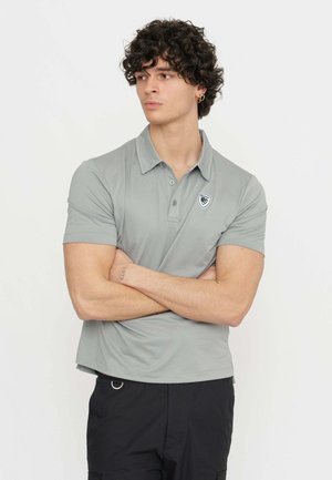 Polo grigia con colletto strutturato, maniche corte e un logo sul petto. Texture liscia, design casual, abbinata a pantaloni corti neri.