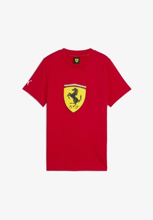 Červené tričko s veľkým žltým erbom Ferrari, na ktorom je čierny skáčúci kôň a „SF“ v strede, doplnené zelenými a bielymi pruhmi.
