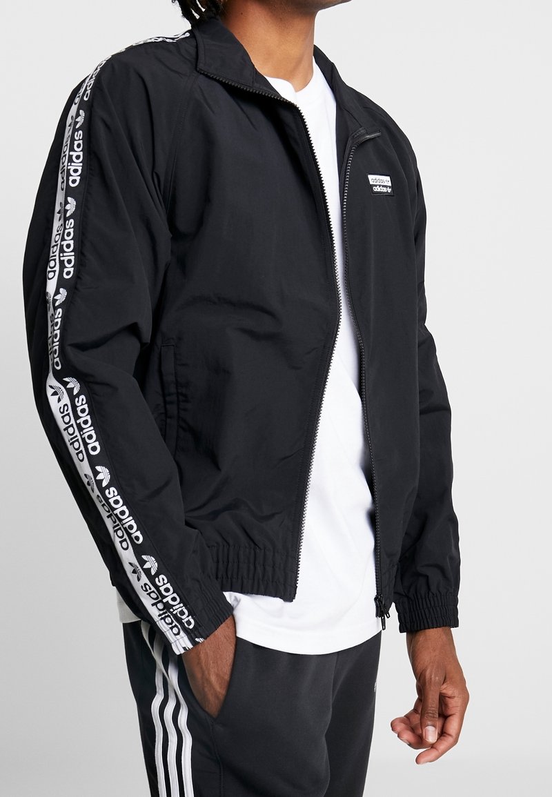 Svart vindjacka med elastiska muddar, sidofickor och dragkedja fram. Har vit Adidas-logoband längs ärmarna.