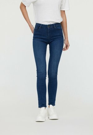 Kvinde iført mørkeblå skinny jeans, hvid T-shirt og hvide platformsneakers, stående foran en ensfarvet lys baggrund.