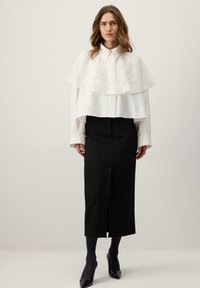 Ženska z valovitimi lasmi, oblečena v belo dolgo srajco z madežem čipke, črno midi krilo, črne nogavice in črne koničaste pete, stoji.