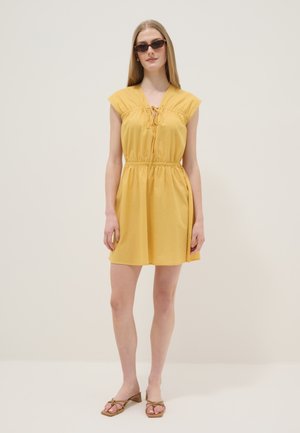 Femme portant une robe jaune sans manches, des sandales marron à talons et des lunettes de soleil, debout devant un fond clair uni.