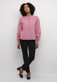 Blouse rose texturée avec encolure ronde et boutonnière, associée à un pantalon noir ajusté et des escarpins à bout fermé, avec une finition lisse.