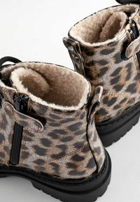 Botas de tobillo con estampado de leopardo y superficie brillante, forro de suave felpa beige y suela de goma negra texturizada; cuentan con cremalleras y tiradores.